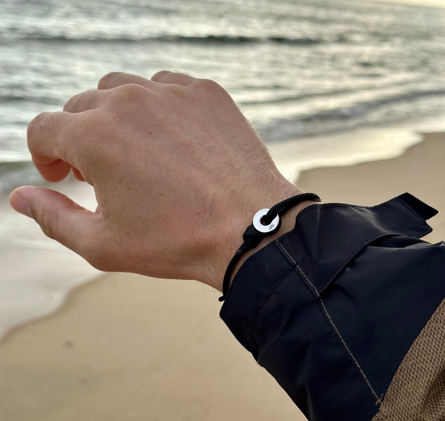 Hand reaching towards the ocean with a sailing bracelet on the wrist, set against a beach backdrop.